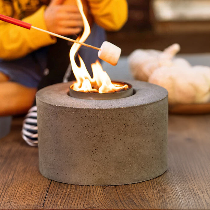 A cylindrical concrete campfire stove with a small fire burning in the center, shown on a wooden floor with a person roasting a marshmallow over it in a camp setting.
